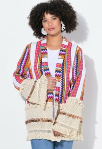 Cardigan intrecciato colorato con motivi geometrici, base bianca, maniche frangiate e accenti decorativi di monete.