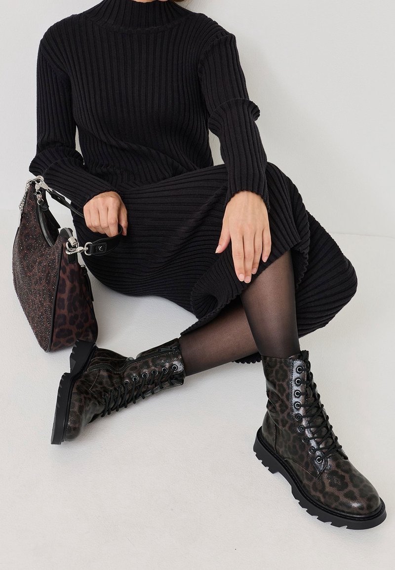 Personne assise portant une robe noire côtelée, des collants noirs transparents, des bottes lacées imprimé léopard, et tenant un sac à main assorti imprimé léopard.