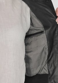 Schwarze Lederjacke mit grauem Innenfutter. Verfügt über eine Reißverschlusstasche und ein sich wiederholendes Logo-Muster. Hell gefärbtes Hemd auf der linken Seite sichtbar.