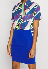 Chemise à manches courtes coupée avec des motifs géométriques multicolores et un logo Vans, associée à une robe bleue ajustée avec des rayures blanches sur les côtés.