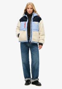 Giacca imbottita con tessuto in shearling blu e crema, dotata di cappuccio, tasche con zip e design testurizzato. Abbinata a jeans blu chiaro.