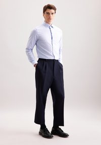 Camicia button-up a quadri azzurro chiaro, pantaloni pleat blu navy scuri e scarpe eleganti nere. Il modello è in piedi con le mani nelle tasche su uno sfondo neutro.