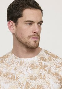 T-shirt blanc avec un motif floral beige, col rond et manches courtes. Le tissu semble lisse avec une texture légère.