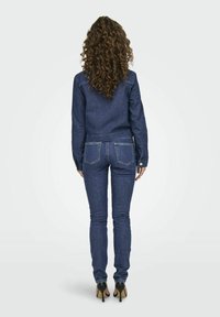 Giacca di denim scuro con maniche lunghe e chiusura con bottoni, abbinata a jeans aderenti in denim scuro. Entrambi presentano cuciture visibili e tasche.