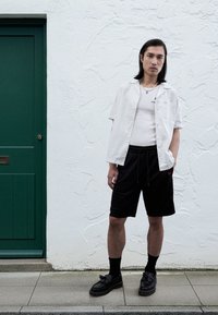 Camicia bianca a maniche corte sopra una canottiera aderente, abbinata a pantaloncini neri con coulisse e mocassini neri. Porta verde sullo sfondo.