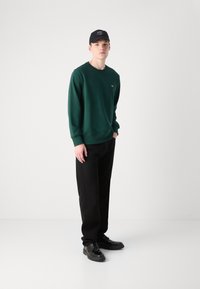 Groene katoenen sweatshirt met geribbelde boorden, gecombineerd met zwarte broek en zwarte schoenen. Een zwarte pet met witte logo-accenten maakt de look compleet.