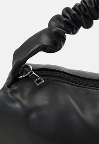 Musta tekonahkainen laukku, jossa on rypytetty kahva ja vetoketjullinen suljin metallisella vetimellä. Sileä pinta, jossa on hienovarainen kiilto.