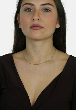 Collana in oro, design sottile con una texture liscia, indossata attorno al collo. La modella indossa una maglietta scura a scollo a V e un trucco minimale.
