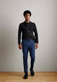 Zwarte lange mouwen polo, op maat gemaakte blauwe geruite broeken, zwarte schoenen en een schoudertas, staande tegen een neutrale achtergrond.