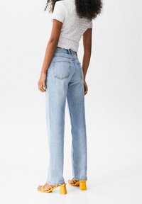 Femme aux cheveux bouclés portant un haut blanc texturé, un jean droit bleu clair et des sandales à talons carrés jaunes, debout de profil.