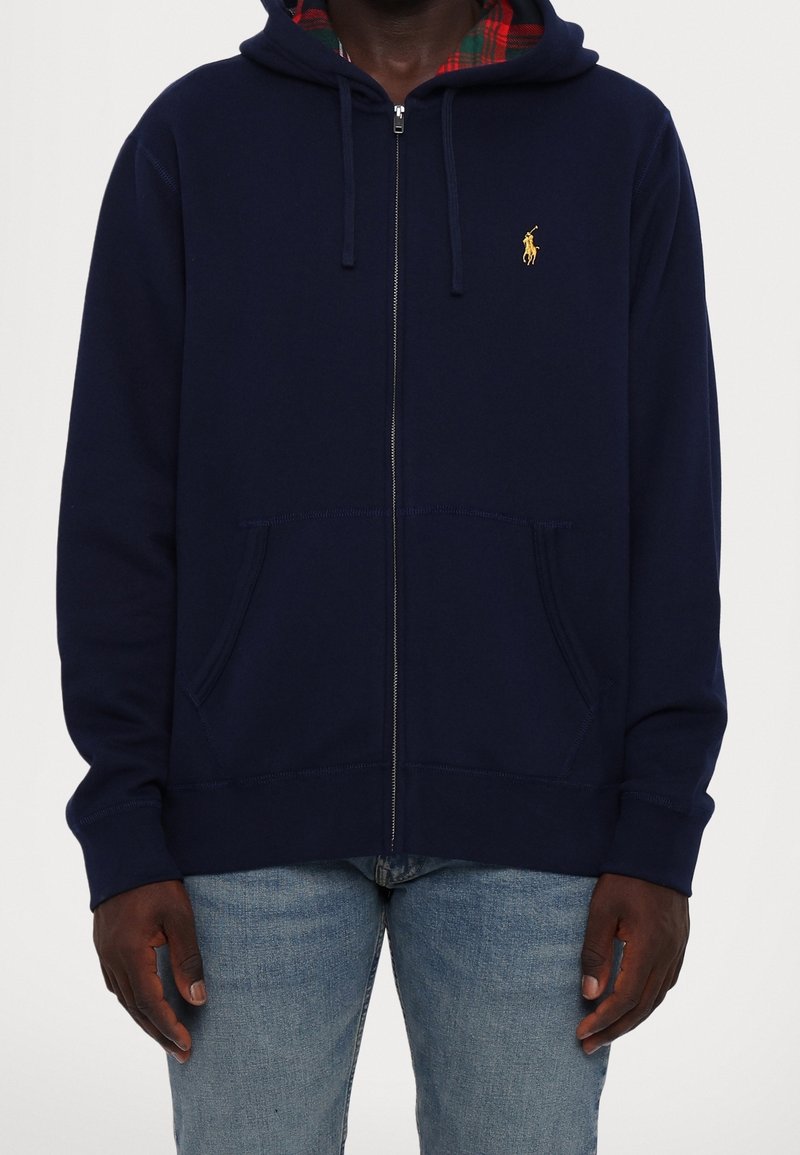 Mężczyzna w granatowej bluzie na zamek z kapturem wyłożonym czerwoną kratą oraz jasnoniebieskich jeansach, z rękami swobodnie opuszczonymi wzdłuż ciała.