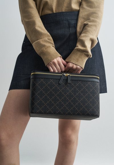 Μαύρη συνθετική τσάντα με γεωμετρικό μοτίβο, χρυσό φερμουάρ και δύο λαβές φερμουάρ. Λείο υλικό, ορθογώνιο σχήμα. Κρατιέται μπροστά από ένα άτομο.