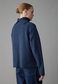 Pull en tricot bleu marine avec col et ourlet côtelés, présentant une texture douce et un design surdimensionné, associé à un jean en denim foncé.