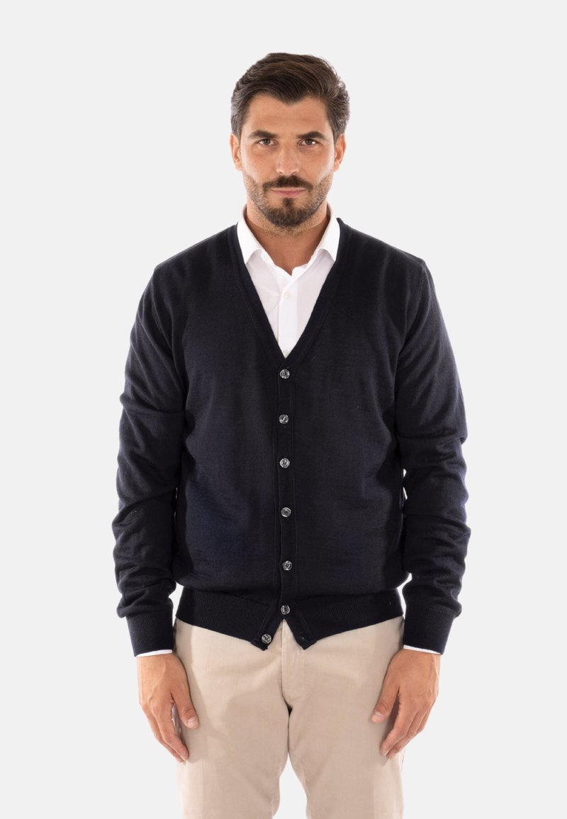 Cardigan in lana blu navy con scollo a V, bottoni frontali, polsini e orlo a costine. Indossato sopra una camicia bianca con colletto, dalla texture liscia.