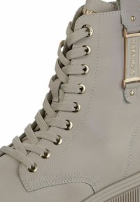 Stivaletto sintetico grigio chiaro con hardware dorato, dotato di lacci piatti, occhielli in metallo e suola in gomma testurizzata.