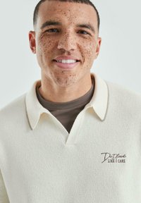 Glunderende man met kort haar en sproeten, gekleed in een crèmekleurig gebreid shirt met een bruine ondershirt en de tekst "Ziet het eruit alsof het me iets scheelt" op de borst.
