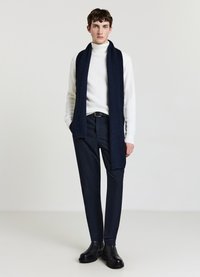 Sciarpa a costine blu navy, dolcevita bianco, pantaloni scuri con motivo e stivaletti neri alla caviglia creano un outfit stratificato con trame diverse.