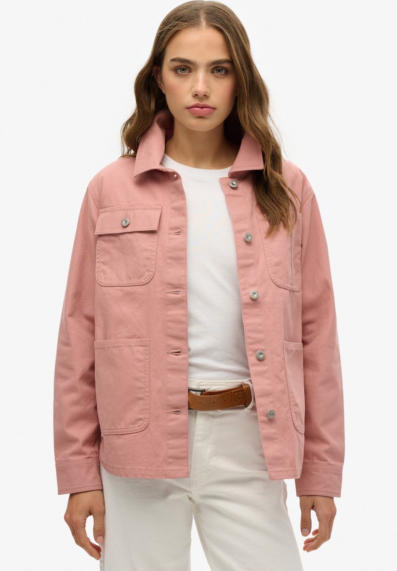 Chaqueta de algodón rosa claro con cuello tipo solapa, botones a presión en la parte delantera, dos bolsillos en el pecho y un corte relajado. Lleva puesta sobre una blusa blanca y pantalones claros.