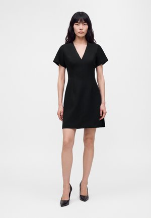 Femme debout portant une robe noire courte à col en V avec des manches courtes et des talons hauts noirs sur un fond blanc uni.