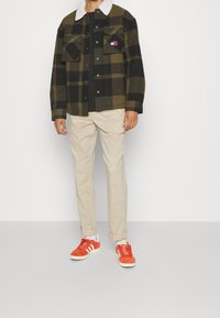 Grön och svart rutig jacka med vit krage, två bröstfickor; beige byxor; orange sneakers med vita detaljer och tre ränder.