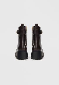 Bottes de cheville en cuir marron avec une semelle noire épaisse, fermeture éclair verticale et une boucle décorative près de la cheville. Texture lisse sur l'ensemble.