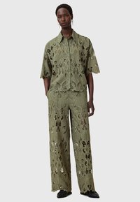 Completo in due pezzi di colore verde oliva composto da una camicia a maniche corte e pantaloni a gamba larga con ritagli in pizzo floreale e bordi ondulati.