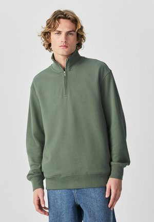 Młody mężczyzna o falowanych blond włosach, ubrany w oliwkowo-zielony sweter z półzamkiem i niebieskie dżinsy, stojący na jednolitym tle.