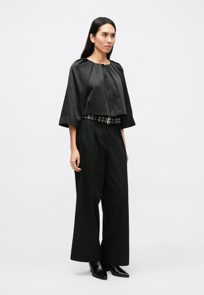 Schwarzes, satinbesetztes Cropped-Top mit Knopfdetail und weiten Ärmeln, kombiniert mit locker sitzenden schwarzen Hosen und einem mit Nieten besetzten Gürtel.