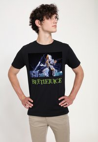 Sort bomulds T-shirt med grafik print fra Beetlejuice, der viser en karakter i stribede klæder, med grøn tekst nedenfor.