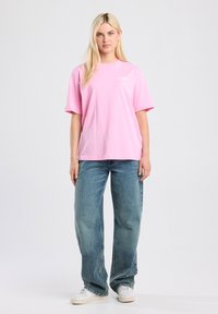 Pink cotton t-shirt with short sleeves, crew neck, and chest logo. Paired with faded blue jeans and white sneakers. Plain background.