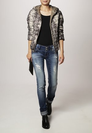 Giacca imbottita con zip, caratterizzata da un motivo a pelle di serpente in beige e marrone, abbinata a jeans blu strappati e stivaletti neri.