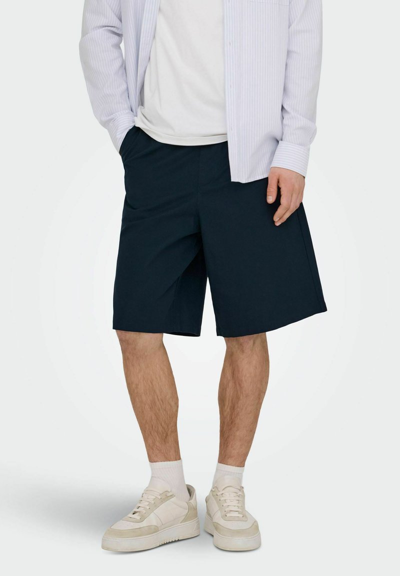 Homme portant un short bleu marine jusqu'aux genoux, des baskets blanches avec des chaussettes et un t-shirt blanc sous une chemise légère à rayures ouverte, les mains dans les poches.