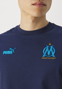 Marinblå bomullströja med blå broderad "PUMA"-logotyp och Marseille-klubbens emblem med guldaccenttext, rund halsringning och korta ärmar.