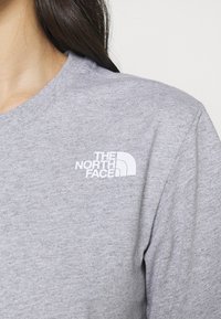 Žena má na sobě světle šedé tričko s kulatým výstřihem a bílým logem The North Face na levé části hrudi.