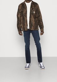 Brun mönstrad jacka över en vit skjorta, tillsammans med blå jeans och vita sneakers med marinblå detaljer. Två bröstfickor på jackan.