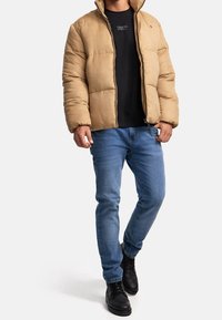 Beżowa puchówka z zamkiem, noszona na czarnej bluzie, w połączeniu z niebieskimi dżinsami i czarnymi sznurowanymi botami. Posiada stójkę.