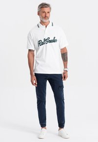 Camisa polo blanca con texto verde "Rad Grader", con un cuello a rayas. Pantalones ajustados azul marino con puños elásticos y bolsillos laterales. Zapatillas blancas.