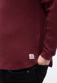 Μπλούζα μακρυμάνικη σε μαρόν χρώμα με ύφανση βαφλοειδούς υφής και ετικέτα Levi's Dry Goods, φορεμένη με μαύρο παντελόνι, με το χέρι τοποθετημένο στην τσέπη του παντελονιού.