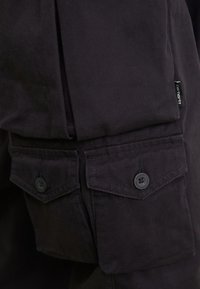 Nahaufnahme von schwarzen Carhartt-Hosen, die mit geknöpften Klappentaschen und einem kleinen Stoffmarkenschild an der Seite versehen sind.