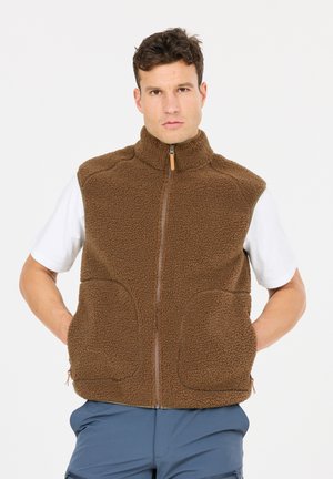 Mann trägt eine braune Fleeceweste mit Reißverschluss über einem weißen kurzärmeligen Hemd und blauen Hosen, steht mit den Händen in den Westentaschen vor weißem Hintergrund.