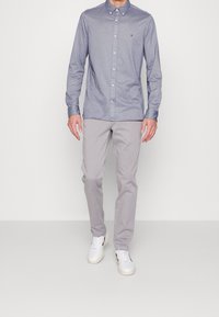 Uomo che indossa una camicia azzurro chiaro con bottoni, pantaloni grigio chiaro e sneakers bianche, mentre cammina su uno sfondo chiaro e uniforme.
