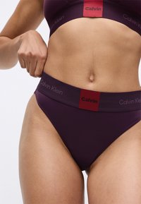 Calvin Klein magas derekú bikinit húzott mélylila színben, kontrasztos piros logós derékrésszel. Sima szövettextúra, minimalista dizájn.