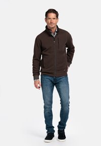 Bruine zip-up jas met gestructureerde stof, zijzakken en borstzak; gecombineerd met blauwe jeans en zwarte schoenen.