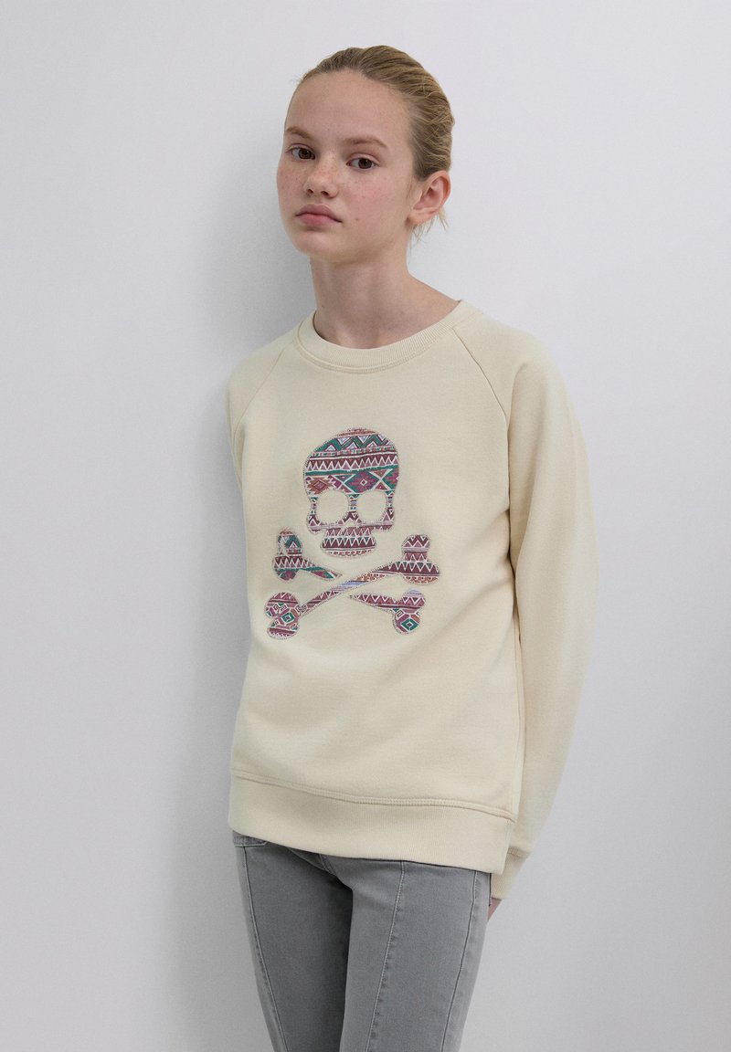 Sweat-shirt beige avec un motif coloré de crâne et os croisés, comprenant des motifs géométriques. Col rond et manches raglan.