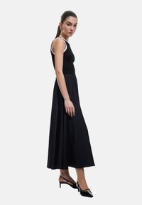 Top de tirantes negro acanalado combinado con una falda larga negra fluida. La modelo lleva zapatos de tacón negros. El fondo es blanco, centrado en el atuendo.