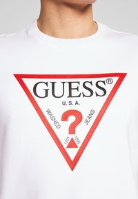Camiseta de algodón blanca con un gran gráfico de triángulo rojo. El texto incluye "GUESS", "U.S.A." y "WASHED JEANS" con un signo de interrogación central.