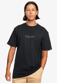 Mustan puuvillan t-paita, jossa on pyöreä kaula-aukko ja edessä keltainen "Quiksilver" -logo. Lyhyet hihat ja rento malli.