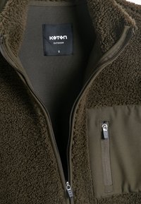 Chaqueta de forro polar verde oliva con cremallera en la parte delantera, tela texturizada y un bolsillo exterior. Presenta una etiqueta de la marca negra en el interior del cuello.