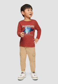 Lange mouwen rode shirt met "FRIENDSAURS" grafiek en blauwe pailletten, gecombineerd met beige cargobroek en witte sneakers.