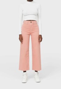 Pantaloni a vita alta e gamba larga in cotone rosa chiaro, con chiusura frontale a bottoni e passanti per cintura, abbinati a un top bianco a maniche lunghe a coste.
